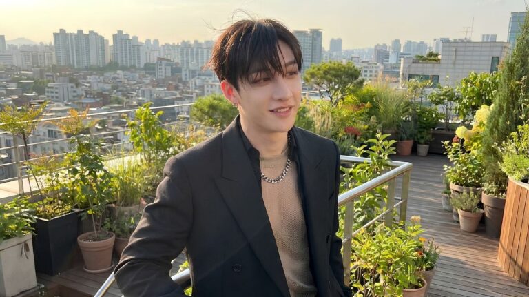 A person in a black suit and mesh shirt stands on a rooftop garden overlooking a city skyline, representing a stylish concept for a blog discussing bangchan age and his evolution as a K-pop leader.